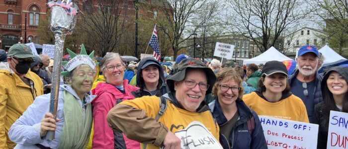 Large group of people outside at a "Hands Off" rally, many in yellow t-shirts, some holding signs.